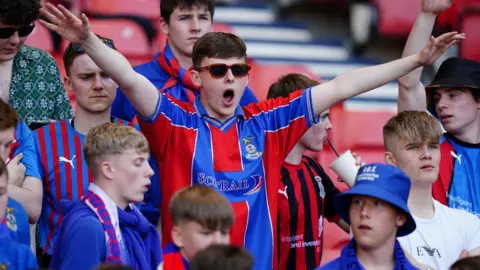 PA Media A group of Inverness Caledonian Thistle fans wearing the club's home kit colours in 2023. One of the fans, who is wearing sunglasses, has his arms raised in celebration.