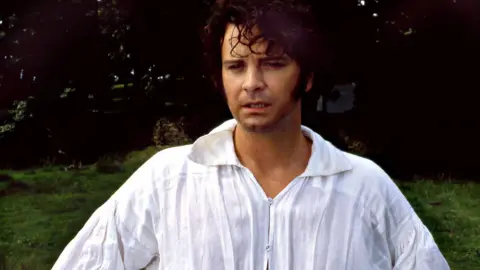 BBC Colin Firth as Mr Darcy in the 1995 BBC drama Pride and Prejudice. He is wearing a white shirt while standing near a pond. Trees can be seen in the backdrop.