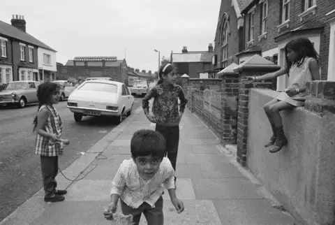 Dennis Morris Black and white photograph of four children hanging around on a residential street.