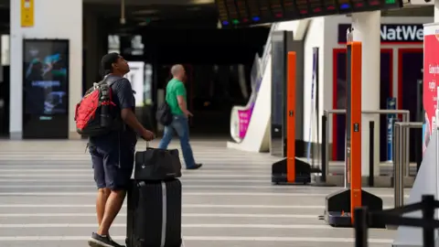 PA Media A passenger looks at message boards at Birmingham New Street Station during strike action in 2022. The man has a case and smaller bag and is carrying a rucksack. Behind him another man wearing a green t-shirt and jeans is walking along. 