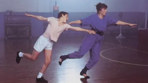 Working Title / BBC Films A scene from the film Billy Elliot shows Jamie Bell as Billy, dancing with Julie Walter, who plays his dance teacher Mrs Wilkinson. They are in a school gym. Billy is wearing a peach-coloured t-shirt, white shorts and socks, and black shoes. He has short dark hair. Mrs Wilkinson is wearing a purple jumpsuit with a black belt tied around her waist. She is wearing black shoes and ankle warmers. Her hair is short and red. 