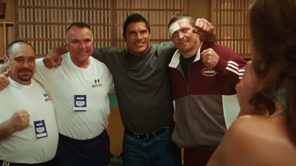 Four men posing together, one wearing an Adidas tracksuit and another in a grey shirt, all smiling and holding fists up.