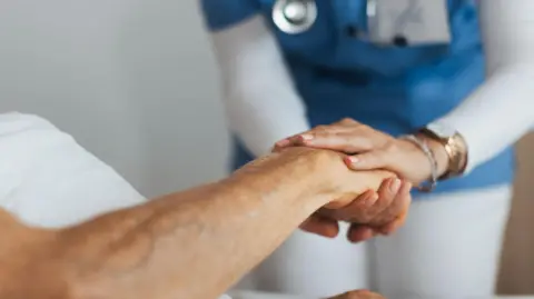 Getty Images A stock photo showing a healthcare professional in blue scrubs holding the hand of a elderly patient lying in a bed.
