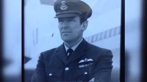 Hansons Auctioneers A black and white photograph of Squadron Leader Denis Lowery. He is wearing his RAF uniform, a hat and with his arms folded.