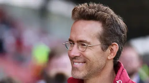 Getty Images Ryan Reynolds smiles and wears red jacket and thin-rimmed glasses