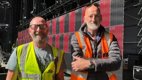 Shivani Chaudhari/BBC Marc and Andy wearing hi-vis jackets on a stage. They are both smiling at the camera. Both have glasses and grey beards.