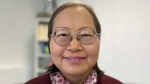 BBC Sophie smiling at the camera. She has brown hair and is wearing round, metal-framed glasses. She is wearing a burgundy cardigan with a pink and white neck scarf.