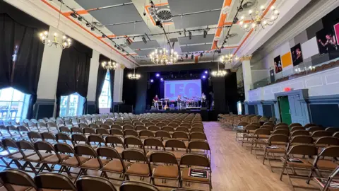 BBC A spacious, elegant hall with rows of brown folding chairs arranged facing a stage. The stage is set up with musical instruments and equipment, and a large screen at the back displays the letters "LEO". The ceiling features ornate chandeliers and acoustic panels, while the walls are adorned with black curtains and framed photographs. The hall is well-lit by natural light coming through tall windows on the left side.