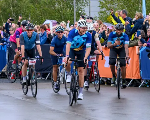 PA/TGI Sport Four cyclists in lycra, helmets and sunglasses, cycling on a smooth road surface, led by Sir Chris Hoy. There are large crowds behind barriers. 