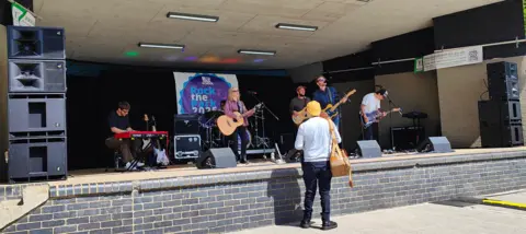 CBSVL A band on a bandstand, with one person standing in front of the structure. There are five musicians on the stage, four with guitars and one on a keyboard. A sign is at the back.