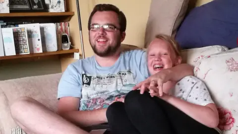 Family handout A man with short brown hair, stubble and rectangular glasses is wearing a blue top and smiling on a sofa with his arm around a teenager. The teenager has her mouth open laughing with braces and has blonde hair tied back, a white top and black leggings. Their hands are entwined. Next to them is a shelf with cards and books on it.