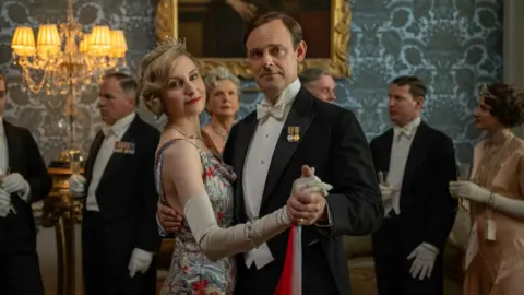 Rory Mulvey 2025 FOCUS Lady Edith stands in her silk floral gown, looking at the camera, with the man who portrays her husband in the film. he is wearing a white shirt and black dinner jacket, they are dancing and looking at the camera.