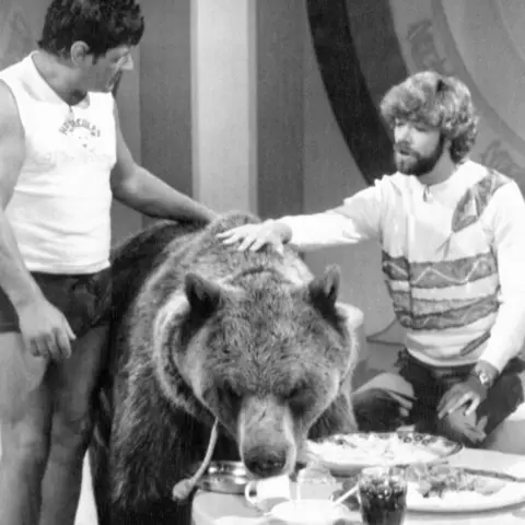 PA Media A black and white photo of two men and a bear on a tv programme. One person stands on the left in casual attire, while the other sits on the right in a patterned shirt. The bear, appearing tame, leans over a table with food.