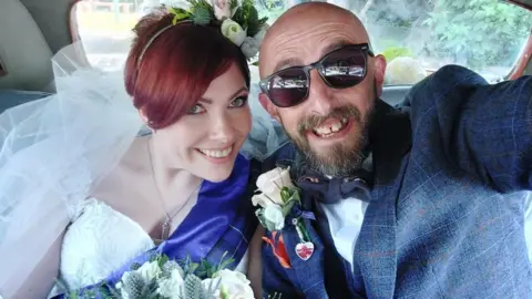 Family photograph Kat Watson-Wood wearing a white wedding dress on her wedding day with husband Matt. They are both smiling in the back of their wedding car after getting married.