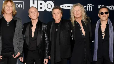 Getty Images Joe Elliott, Phil Collen, Rick Allen, Rick Savage and Vivian Campbell of Def Leppard attend the 2019 Rock & Roll Hall Of Fame Induction Ceremony. There are logos behind them on the backdrop. The men are dressed in black and Vivian Campbell has a grey scarf on.