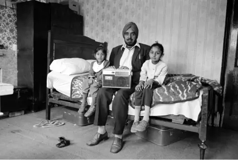 Dennis Morris Black and white photograph of a man sitting on a bed with his two small daughters sat on either side of him. A radio set is perched on the man’s lap