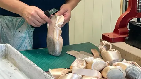 BBC A pile of worn pink pointe ballet shoes sits on a desk. Behind them Rachel holds a knife and is cutting the leather out of the shoes.