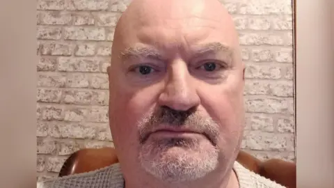 Rich Mulligan A selfie of a man with a grey beard and bald head against a white-washed brick wall.