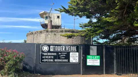 Richard Smith/BBC The circular brick fort is pictured behind black hoarding. It has a small wooden watchtower on top and a large tree to the right. On the black hoarding, which is preventing access, is a green "to let" sign and another black sign advertising it as a wedding venue.