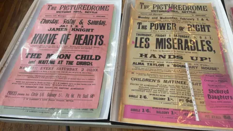 BBC Two cinema posters from the 1920s advertising films at 'The Picturedrome' at Victoria Hall in Settle.
One of the posters is pink whilst the other is yellow. They are both protected by a plastic jacket.
The pink poster is advertising several productions called 'Knave of Hearts', 'The Moon Child' and 'Waiting at the Church'.
The yellow poster is advertising 'The Power of Right', 'Les Miserables' and 'Hands Up!'.