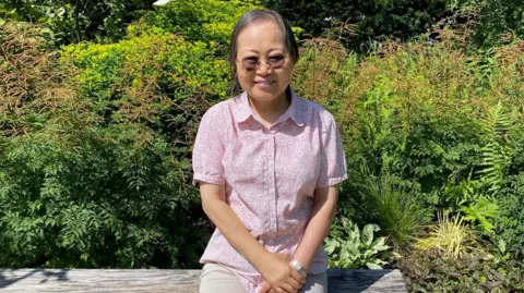 Maggie's Sophie, sat in a sunny garden at Maggie's in Manchester. She is wearing sunglasses, light-coloured trousers and a short-sleeved pink shirt. She is sat on a wooden bench and there are plants and shrubs behind her.