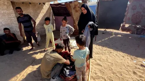 A man crouches down on a sandy floor, and rummages through a bag in al-Mawasi. He is wearing a brown t-shirt and the back of his head is visible. Watching him are three children, a woman and two men nearby. The group stands near a brick wall and some makeshift dividers.