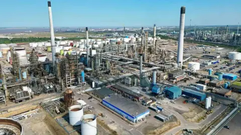 BBC Aerial view of the Lindsey Oil Refinery, a large industrial complex with numerous tall chimneys, large cylindrical storage tanks and a network of interconnected pipelines. Various buildings can be seen throughout the facility, which is surrounded by roads and open land beneath a clear blue sky.