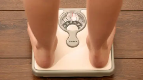 PA Media A pair of bare feet on white scales. The scales are on a brown wooden floor.