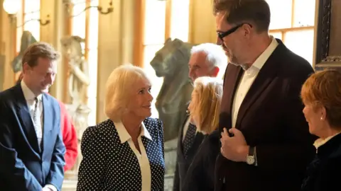 PA Media Queen Camilla meets author Richard Osman at a reception for literary figures during The Queen's Reading Room Festival at Chatsworth House. He is very tall and Queen Camilla is having to look up at him to talk.