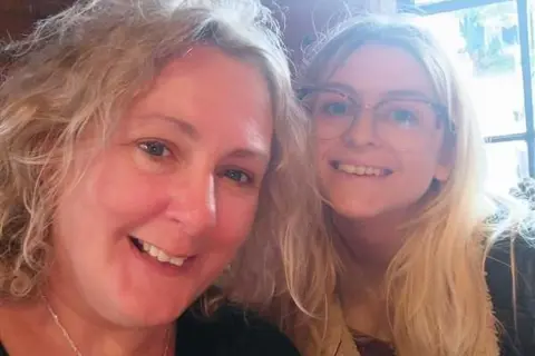 Sarah Gascoyne A mother taking a selfie with her adult daughter. The mum has curly blonde hair and is smiling on the left of the frame. Her daughter, also smiling, has long blonde hair and wears wide-rimmed glasses. They are standing in front of a window with light streaming in.