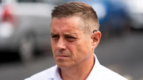 PA Media John Alford in a white shirt, looking to his right. He is clean shaven. He has short dark, greying hair, gelled into a quiff.