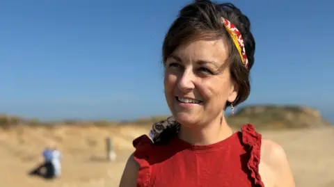 Sarah Wheldon A smiling Sarah Wheldon head and shoulders. She is standing on a beach on a sunny day wearing a red high-neck sleeveless top with a frill trim around the arms. She has brown hair that reaches her neck and a red, white and yellow tie-died scarf tied as a headband.