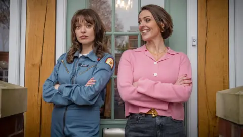 BBC / Gaumont / Ben Blackall Aimee Lou Wood and Suranne Jones stand in front of a green front door, both with their arms crossed, dressed as characters Evie and Suz in a publicity still from Film Club. Wood looks directly at the camera with a straight face while Jones smiles and looks at Wood.