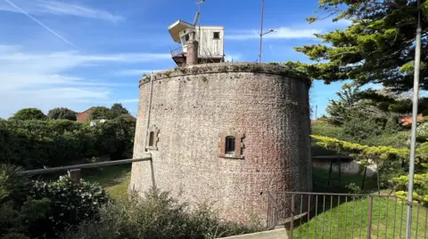 Richard Smith/BBC The 10m (33ft) tall tower is made of brick and surrounded by a dry moat. There are also bushes and trees around the moat. The structure has two small windows and a watchtower on top.