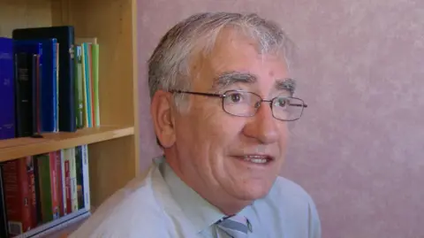 BBC A grey-haired man with glasses, wearing a grey shirt and striped tie, looks beyond the camera. In the background is a bookcase