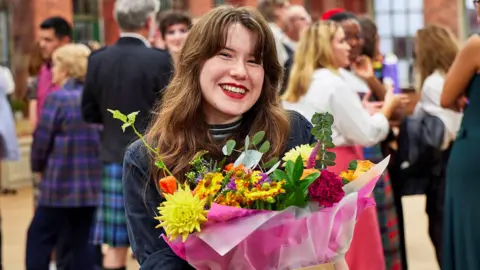 BBC/Love Productions Ltd Órla is standing on The Great British Sewing Bee set. She has long, wavy brown hair and is wearing a dark blue shirt over a stripy top. She is holding a bunch of flowers.