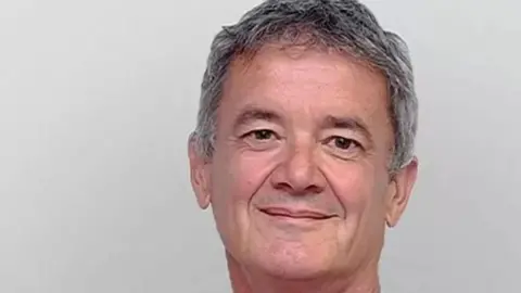 Tony Dixon smiling with short grey hair and brown eyes with grey background.