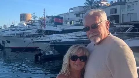 Gaynor Loguello A woman with long blonde hair and wearing sunglasses and a sundress standing with a man who has short grey hair and a grey beard and is wearing a beige t-shirt. They are stood on a harbour with a number of yachts moored in the background.