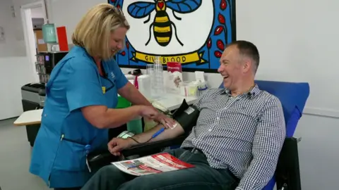 BBC Roger Johnson, who has short hair and is sitting on a blue chair in a medical room, is giving blood. A nurse with blonde bobbed hair and wearing a light blue uniform is collecting blood from Roger's arm. They are both smiling.