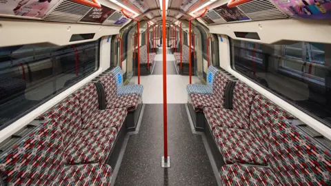 TfL The inside of a Central Line tube carriage. Patterned red and blue seating runs along either side of the carriage, with windows above showing darkness outside. Red poles run from floor to ceiling in the middle of the carriage seating area, and in the standing areas by the doors.