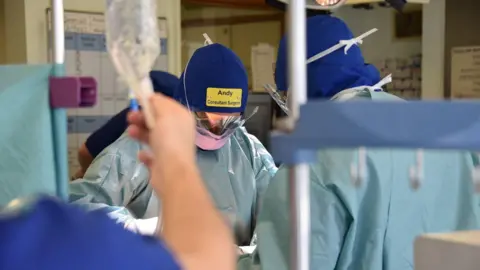 Somerset NHS Foundation Trust A man wearing medical scrubs appears to be performing surgery. On his blue hat, he is wearing a yellow badge which says 'Andy Consultant Surgeon'. He is opposite a colleague, and in the foreground, someone is holding up an IV drip.