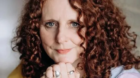 Dasha Tenditna Maggie O'Farrell sits with her hand clasped together under her chin. She has red curly hair and blue eyes.