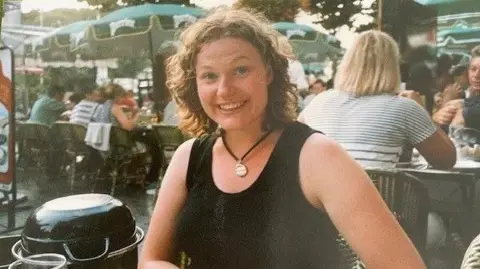 Monica Chatterton A woman sits at a table in a Parisian outdoor cafe and smiles for the camera. She is surrounded by people seated at other tables. She has curly brown hair and wears a black sleeveless top and a gold pendant around her neck.