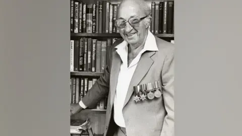 Supplied Alfonso Garizio is pictured in a black and white photograph. He is stood in front of a book case containing several books. He is wearing a grey suit jacket with white open-necked shirt underneath, and has four military medals attached to his left chest. He wears large-framed glasses and is smiling at the camera