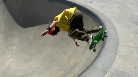 BBC A man skating in a concrete bowl at a skatepark. He is wearing a yellow t shirt and green khaki trousers