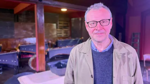 Lee Hall is smiling slightly at the camera. He has short grey hair and wears black-rimmed glasses, beige coat and a grey jumper over a grey shirt. 