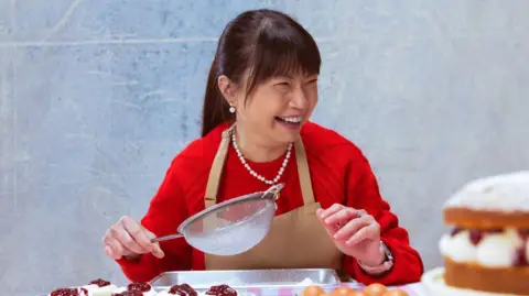 Channel 4/Love Productions Pui Man is wearing a brown apron and a red jumper. She is holding a sieve with cupcakes and eggs on a table in front of her. She has black hair with a fringe and in a ponytail and the back and is laughing.