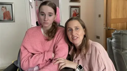 Rachel Cowley A young girl in a pink jumper in a wheelchair. Her mum is sat next to her on the right of the image wearing a pink top and has a smart watch on. Behind them you can see photos of other family members on the walls.