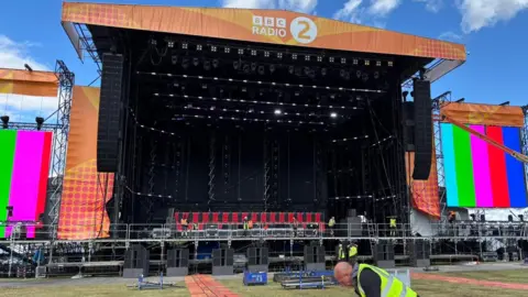 Shivani Chaudhari/BBC An outdoor music stage with a Radio 2 banner on it and TV screens on the sides. It is being set up, with various workers in hi-vis jackets on stage.
