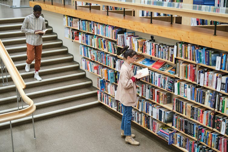 A woman reads a book by a library bookshelf